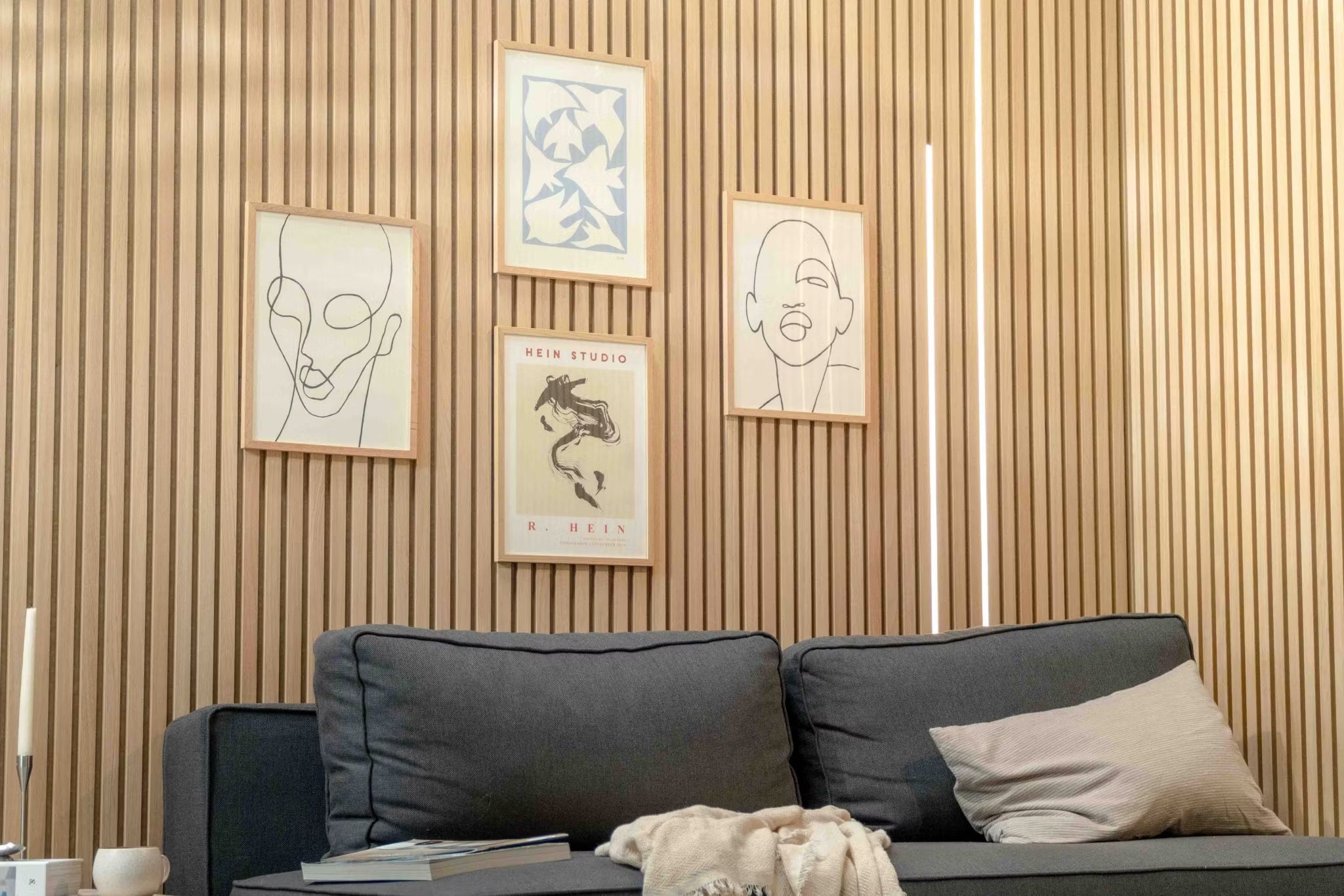 Soggiorno moderno caratterizzato da un divano scuro con cuscino beige, accentuato da una parete di doghe acustiche in legno Akupanel nordico e da unarte scandinava minimalista in alto.